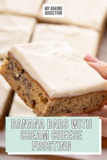 Hand holding a frosted banana bar up to the camera; the pan of sliced bars is visible in the background. Text overlay includes recipe name.