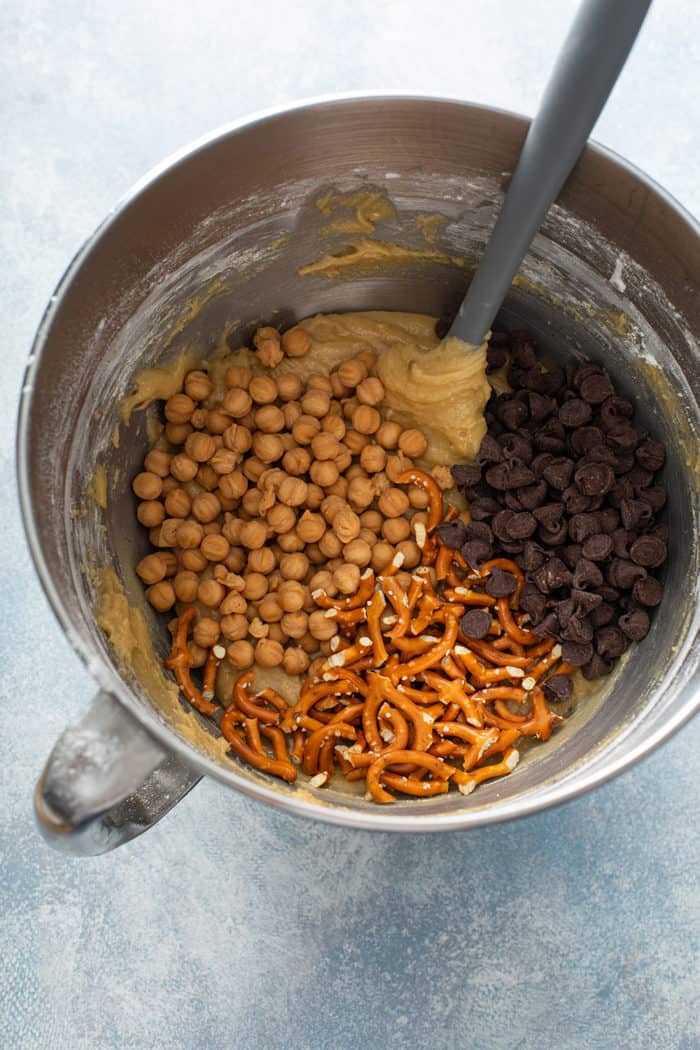 Caramel bits, chocolate chips, and pretzels being added to blondie batter in a metal mixing bowl