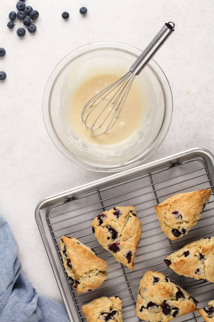 Baked blueberry scones cooling on a wire rack next to a bowl of lemon glaze.