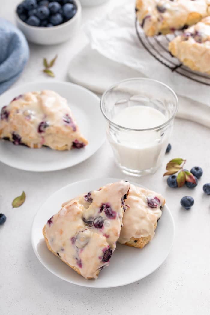 Two white plates with glazed blueberry scones next to a glass of milk.