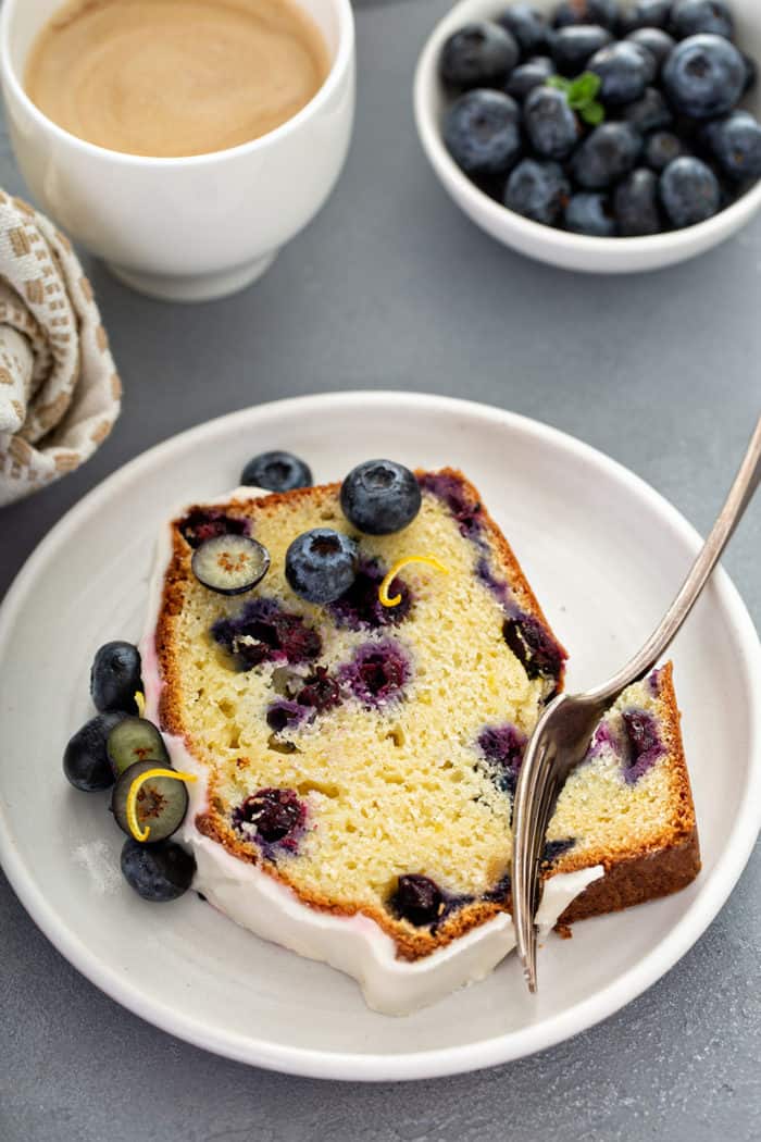 Fork taking a bite out of the corner of a piece of lemon blueberry bread on a white plate