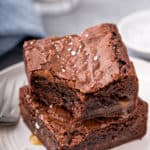 Two salted caramel brownies stacked on a white plate. The top brownie has a bite taken out of it and caramel is pooling out of the edges of the brownies