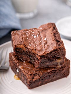 Two salted caramel brownies stacked on a white plate. The top brownie has a bite taken out of it and caramel is pooling out of the edges of the brownies