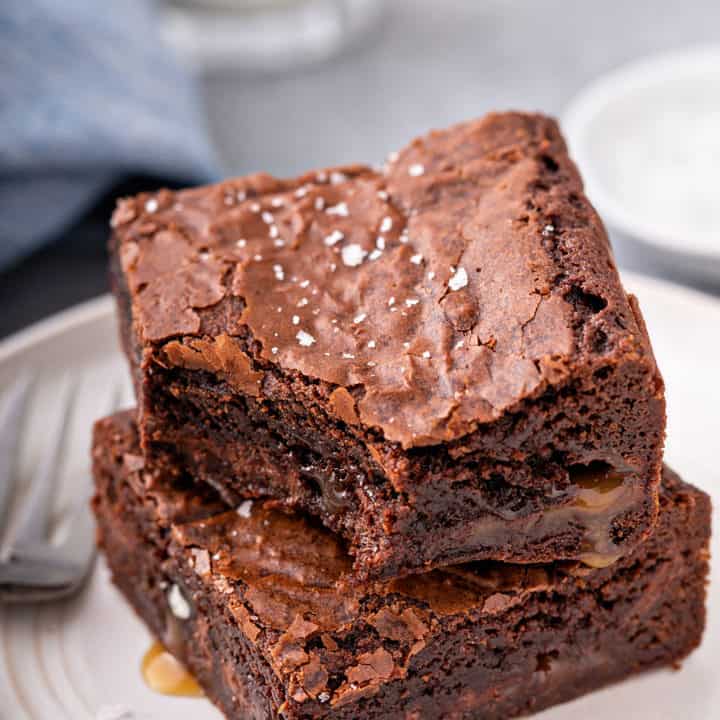 Two salted caramel brownies stacked on a white plate. The top brownie has a bite taken out of it and caramel is pooling out of the edges of the brownies