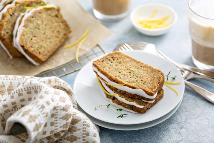 Two slices of lemon zucchini bread stacked on a white plate, with additional slices of the bread and a cup of iced coffee in the background