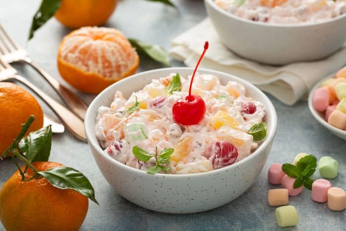 Ambrosia salad topped with a maraschino cherry in a small white bowl, surrounded by mandarin oranges and mini marshmallows