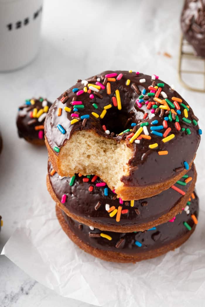 Three chocolate glazed donuts stacked on a piece of parchment paper. The top donut has a bite taken out of it.