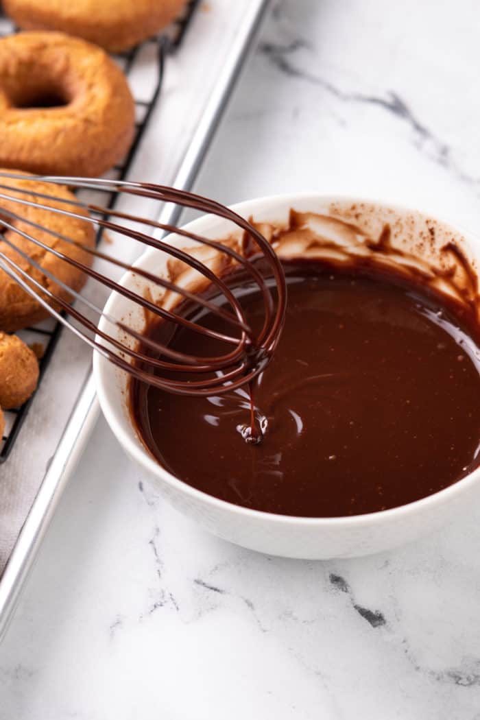 Whisk stirring chocolate glaze in a white bowl next to a pan of donuts.