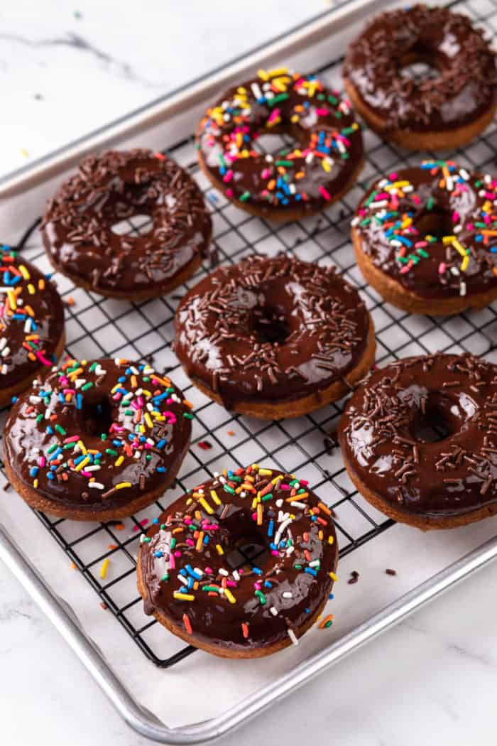 Chocolate glazed donuts, topped with sprinkles, lined up on a wire cooling rack on top of a baking sheet.