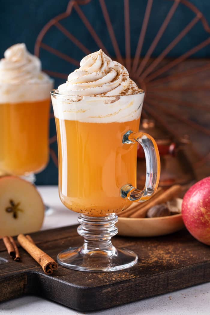 Close up of glass mug filled with spiced apple cider. The cider is topped with whipped cream and dusted with ground cinnamon.