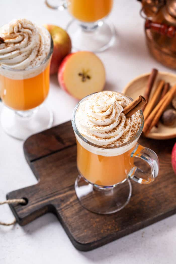 45-degree overhead view of a glass mug of spiced apple cider, topped with whipped cream and garnished with a cinnamon stick.