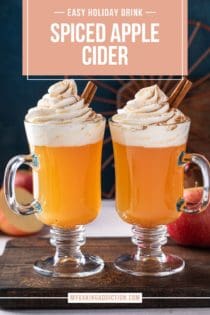 Two glass mugs filled with spiced apple cider and garnished with whipped cream and cinnamon sticks. Text overlay includes recipe name.