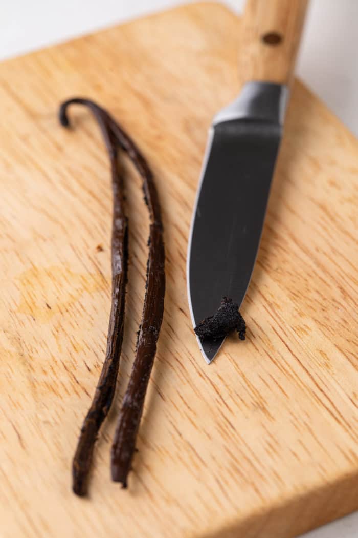 Split vanilla bean next to a paring knife on a wooden cutting board.