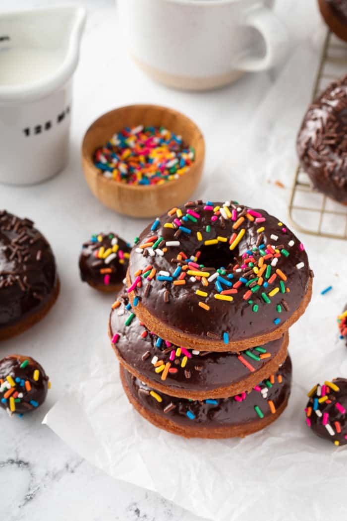 Three sprinkle-topped chocolate glazed donuts stacked on a piece of parchment paper, with more donuts and sprinkles in the background.