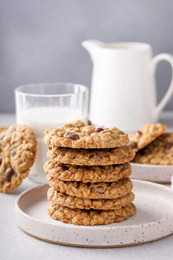 Six oatmeal raisin cookies stacked on a white plate in front of a glass of milk.