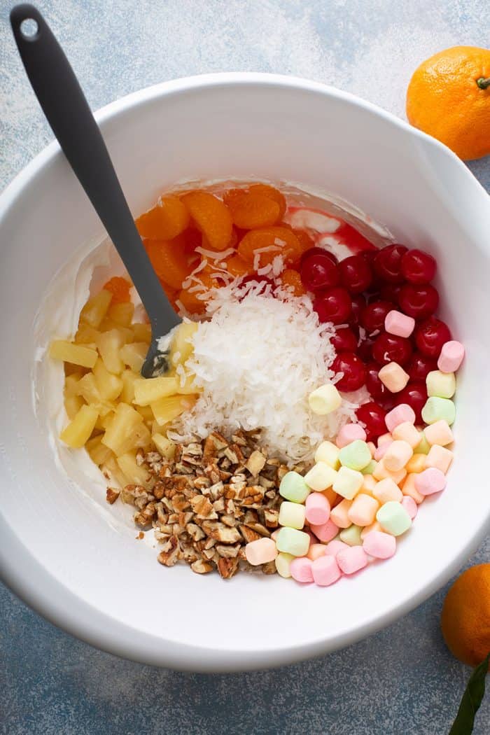 Spatula folding in fruit, marshmallows, coconut, and nuts into a white mixing bowl