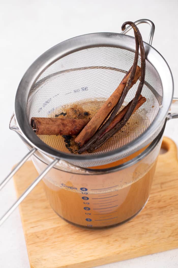Strained spiced apple cider in a glass measuring cup, with the strainer filled with cinnamon sticks set on top.