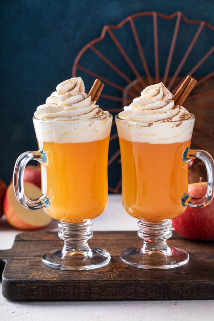 Two glass mugs filled with spiced apple cider and garnished with whipped cream and cinnamon sticks.