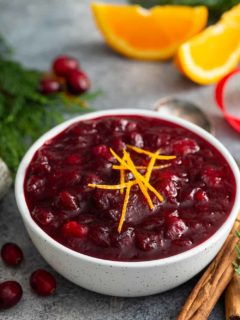 Cranberry sauce in a white bowl, surrounded by greenery and cinnamon sticks