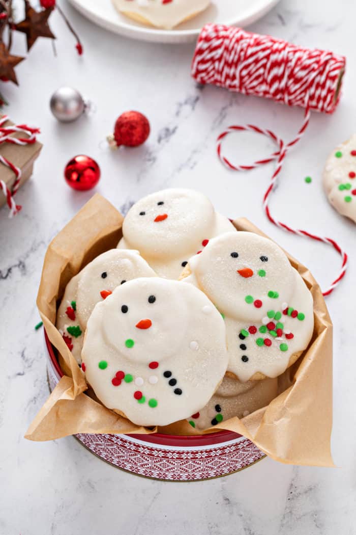 Melted snowman cookies in a festive holiday tin, set on a marble countertop.