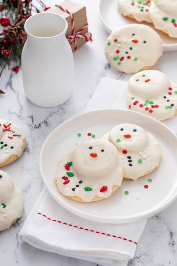 Two melted snowman cookies on a white plate with a glass of milk in the background.
