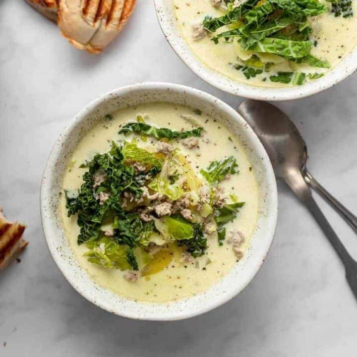 Zuppa toscana in white bowls on a marble countertop next to spoons and bread