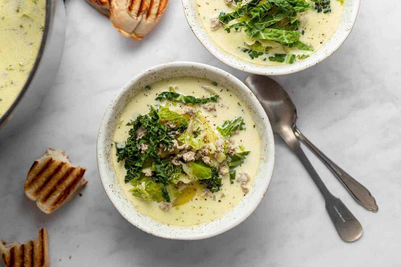 Zuppa toscana in white bowls on a marble countertop next to spoons and bread