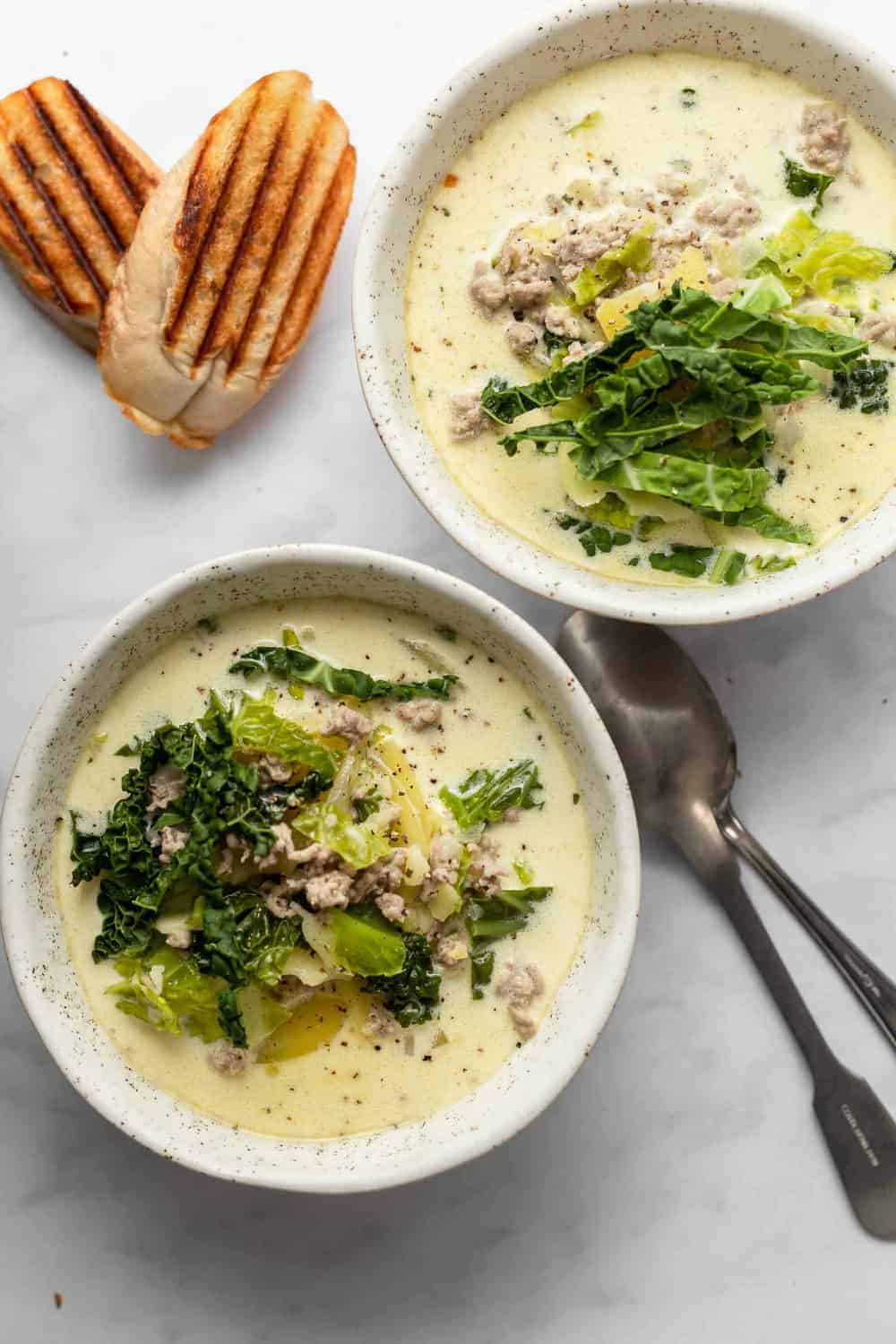 Two white bowls filled with zuppa toscana on a marble countertop next to grilled bread