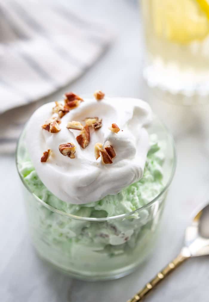 Close up of watergate salad in a small serving bowl, topped with whipped topping and chopped pecans, next to a spoon