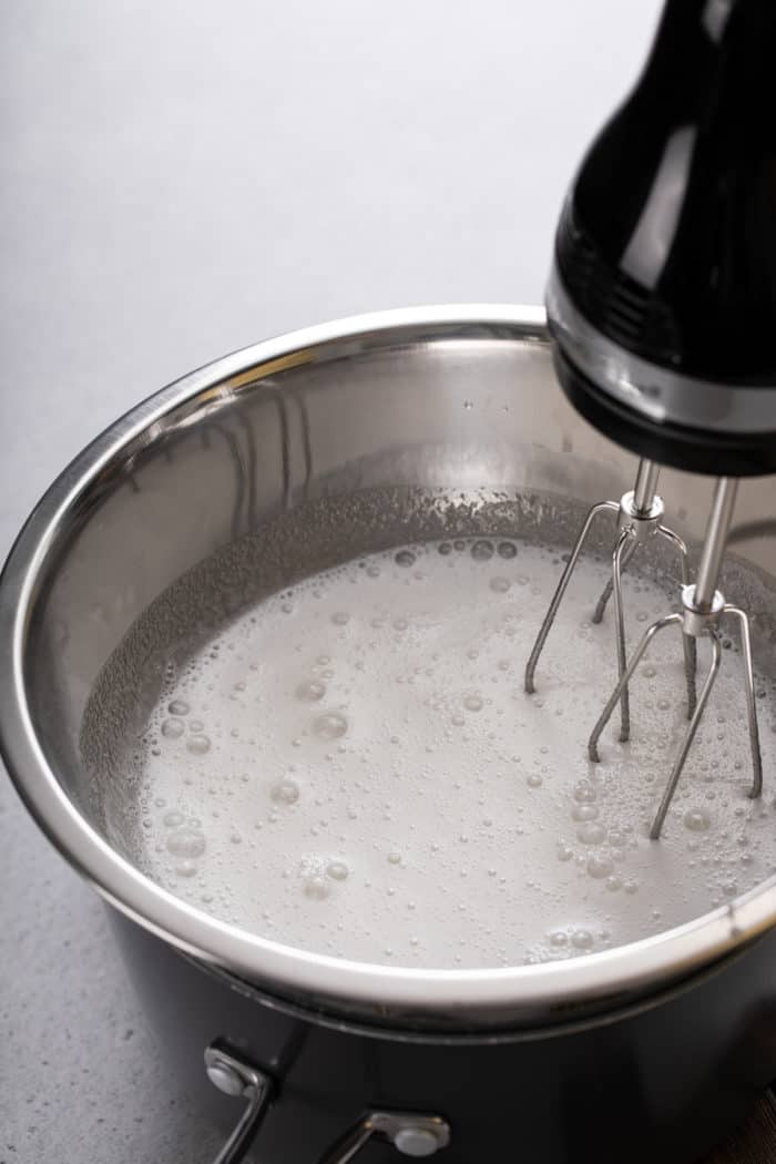 Hand mixer mixing marshmallow frosting in a metal bowl over boiling water.