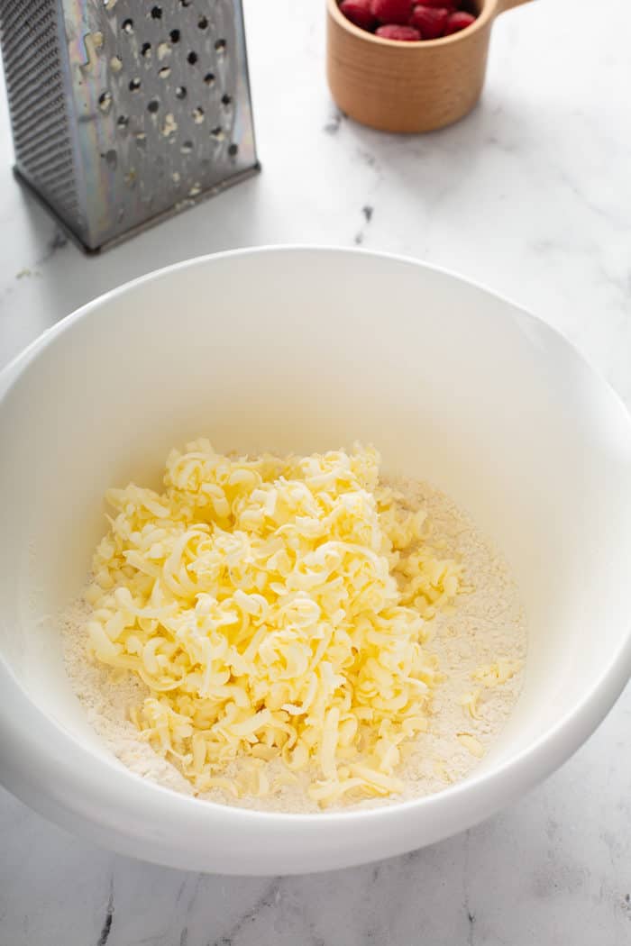 Grated butter added on top of the dry ingredients for scones in a white mixing bowl