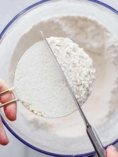 Hand holding a brass measuring cup over a bowl of flour while a knife smooths out the top