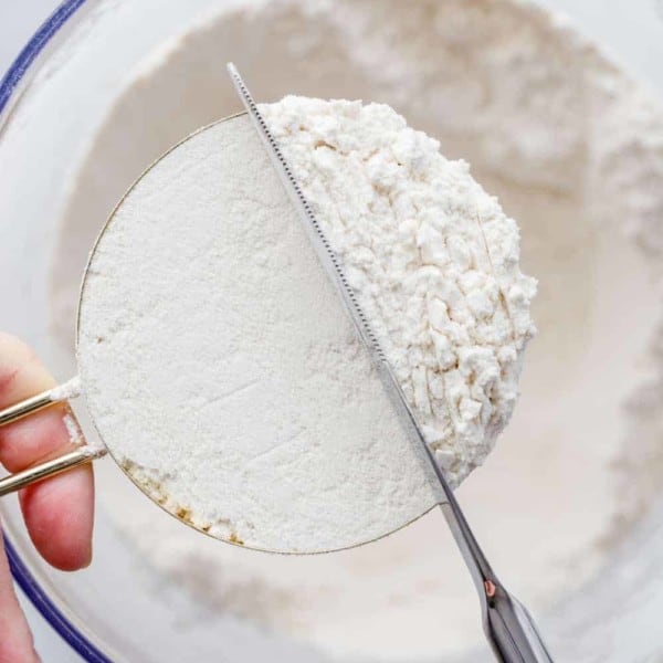 Hand holding a brass measuring cup over a bowl of flour while a knife smooths out the top