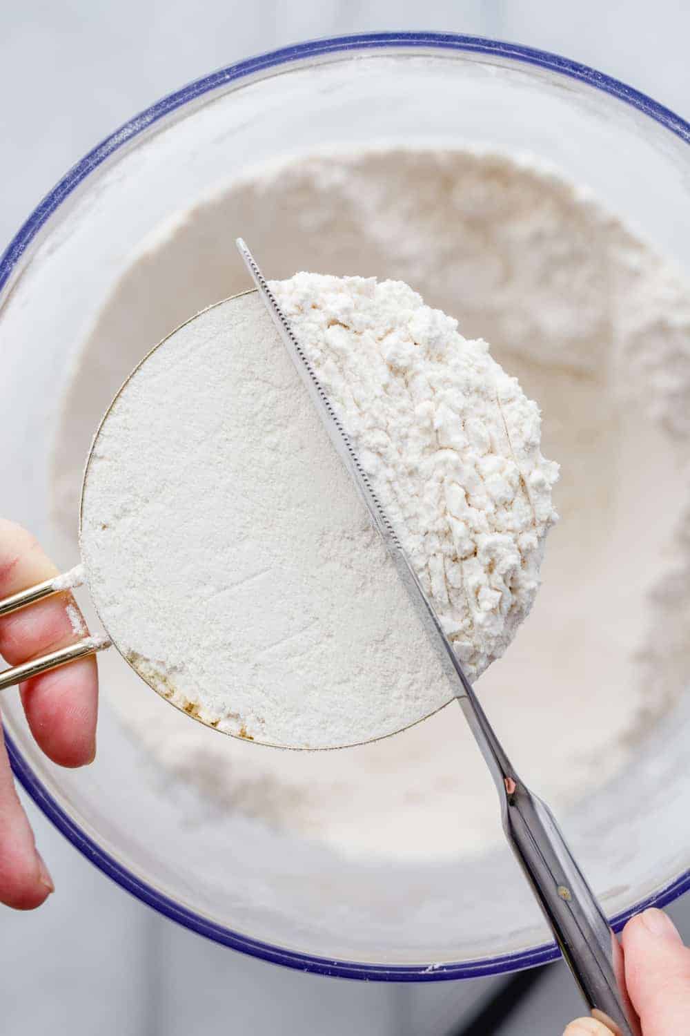 Hand holding a brass measuring cup over a bowl of flour while a knife smooths out the top