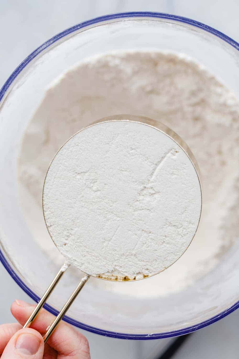 Hand holding a level measuring cup of flour over a bowl of flour