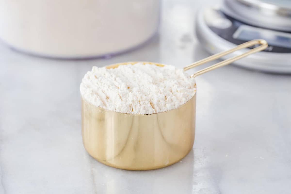 Gold measuring cup full of unleveled flour on a white surface