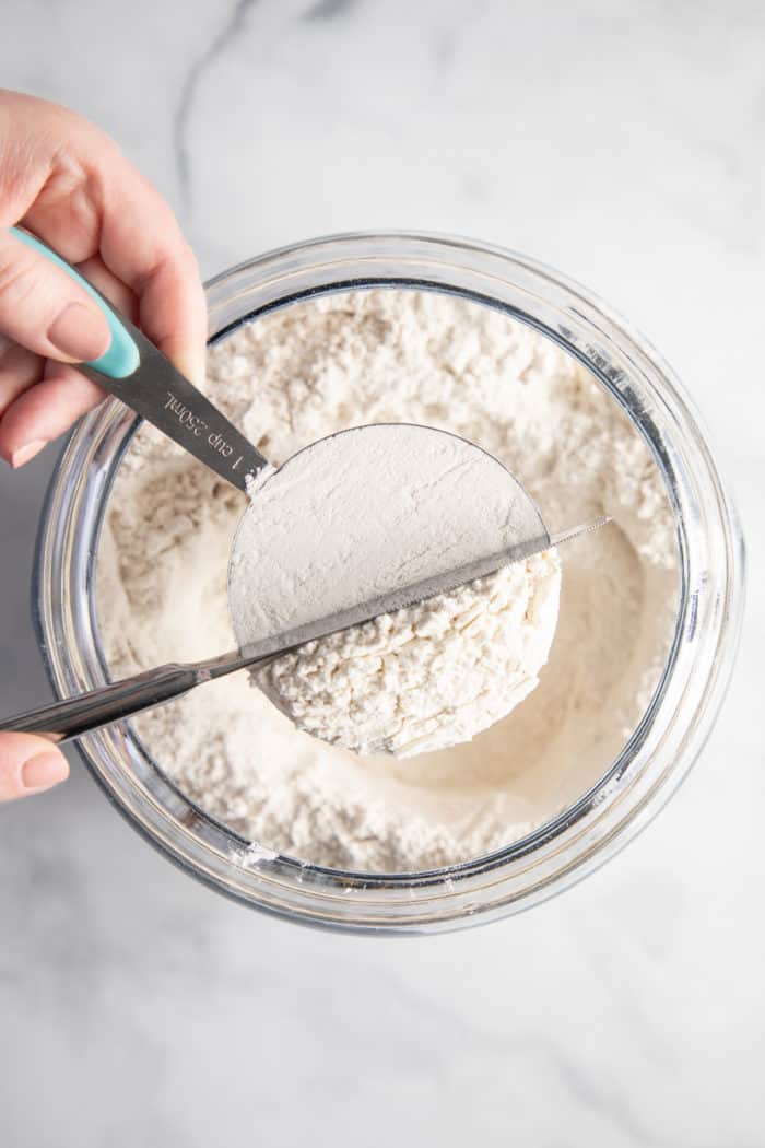 Hand using a knife to level off the top of a measuring cup of flour over a bowl.