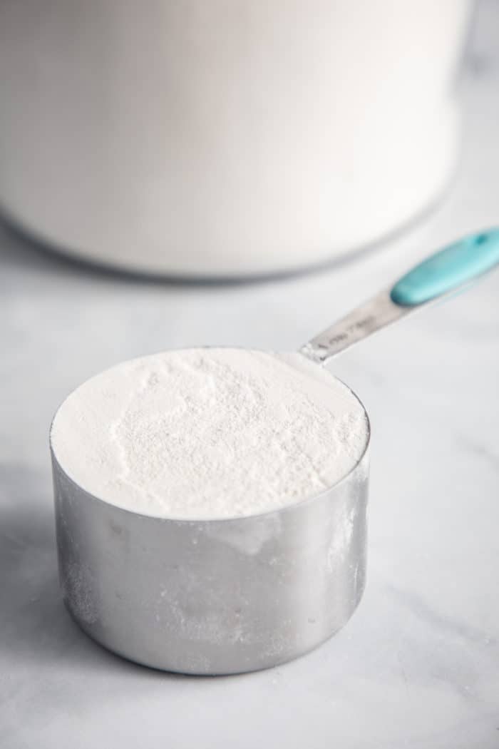 Leveled measuring cup of flour set on a marble countertop in front of a canister of flour.