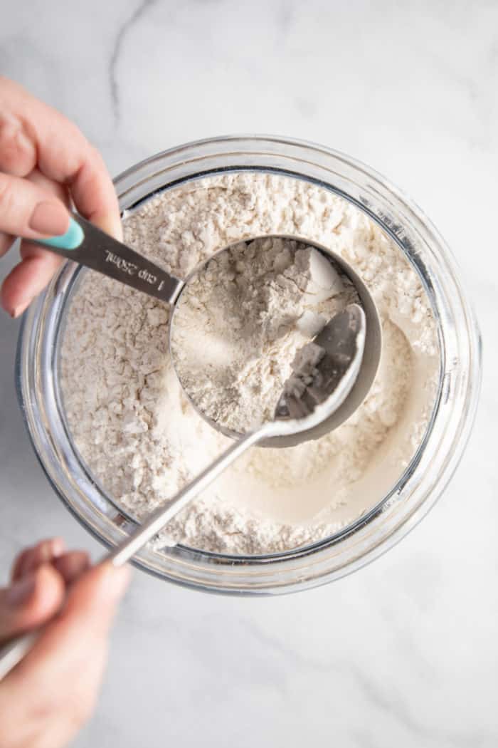 Hand spooning flour into a measuring cup held over a bowl of flour.
