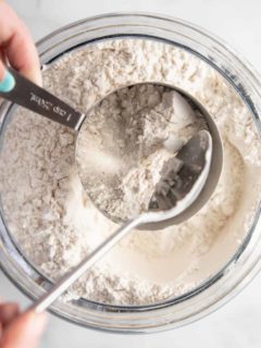 Flour being spooned into a metal measuring cup over a bowl of flour.