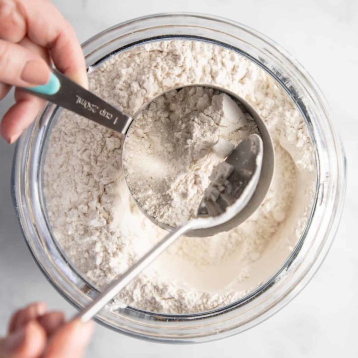Flour being spooned into a metal measuring cup over a bowl of flour.