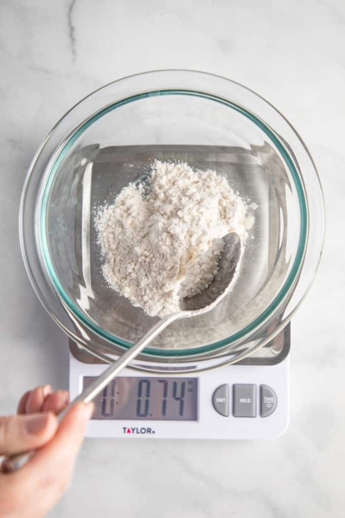 Flour being spooned into a glass bowl set on a kitchen scale.