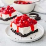 Slice of no bake oreo cheesecake topped with cherry pie filling on a white plate with a 2nd slice and a bowl of cherry pie filling in the background