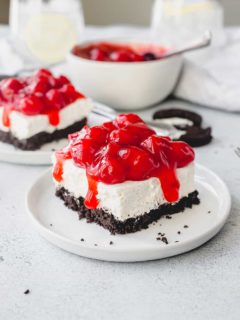 Slice of no bake oreo cheesecake topped with cherry pie filling on a white plate with a 2nd slice and a bowl of cherry pie filling in the background