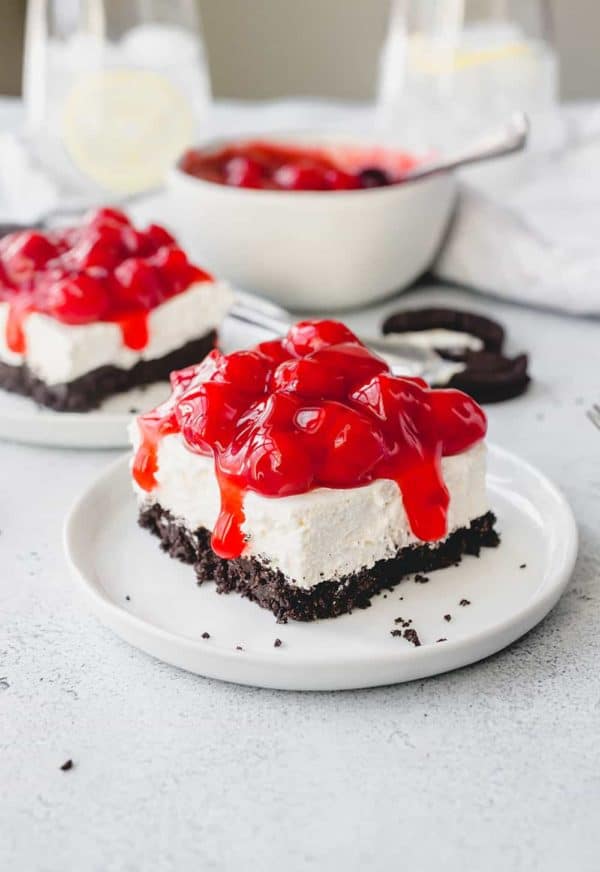 Slice of no bake oreo cheesecake topped with cherry pie filling on a white plate with a 2nd slice and a bowl of cherry pie filling in the background
