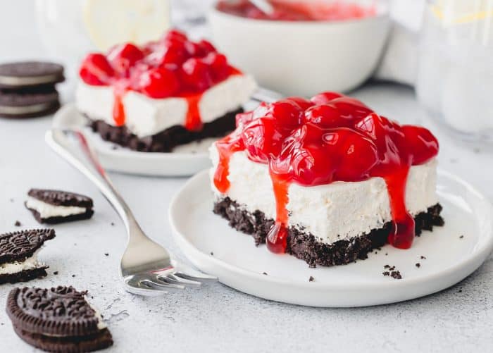 Two slices of no bake oreo cheesecake on white plates, topped with cherry pie filling