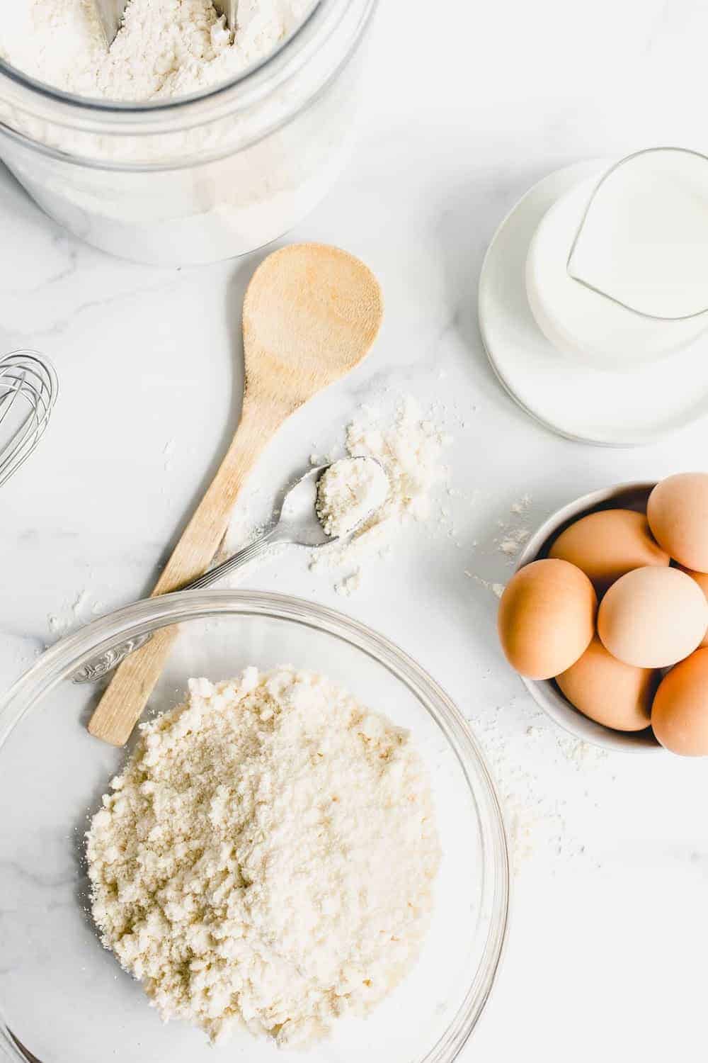 Homemade bisquick mix in a glass mixing bowl on a marble surface surrounded by milk and eggs