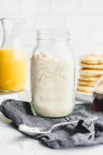 Homemade Bisquick mix in a glass jar with a stack of pancakes and a carafe of orange juice in the background