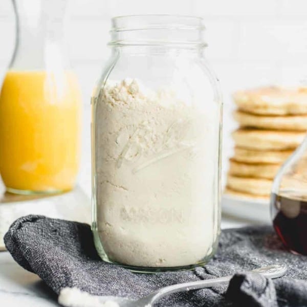 Homemade Bisquick mix in a glass jar with a stack of pancakes and a carafe of orange juice in the background