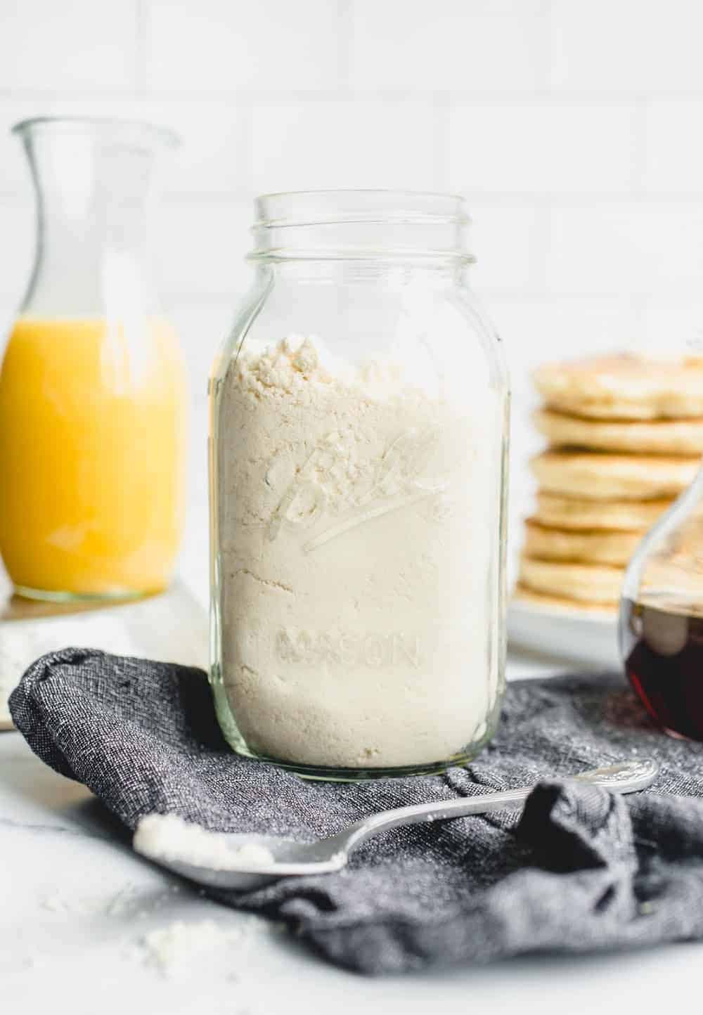 Homemade Bisquick mix in a glass jar with a stack of pancakes and a carafe of orange juice in the background